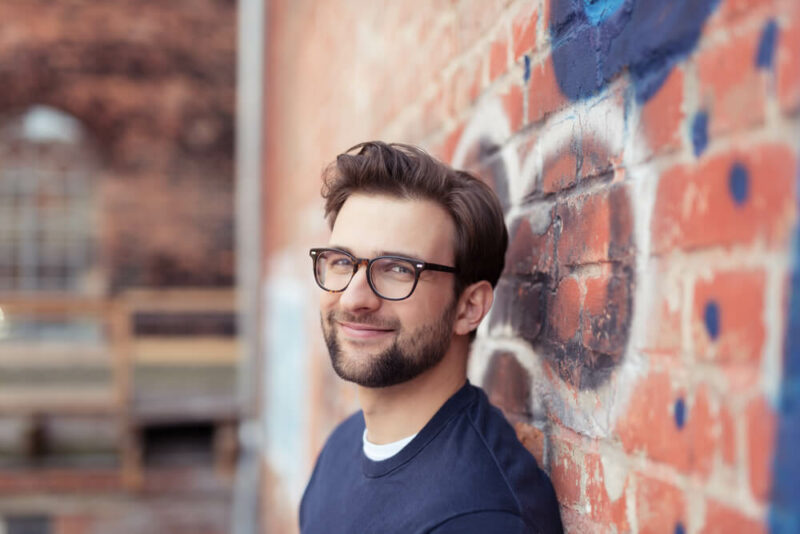 Smiling young man with glasses leaning against a brick wall